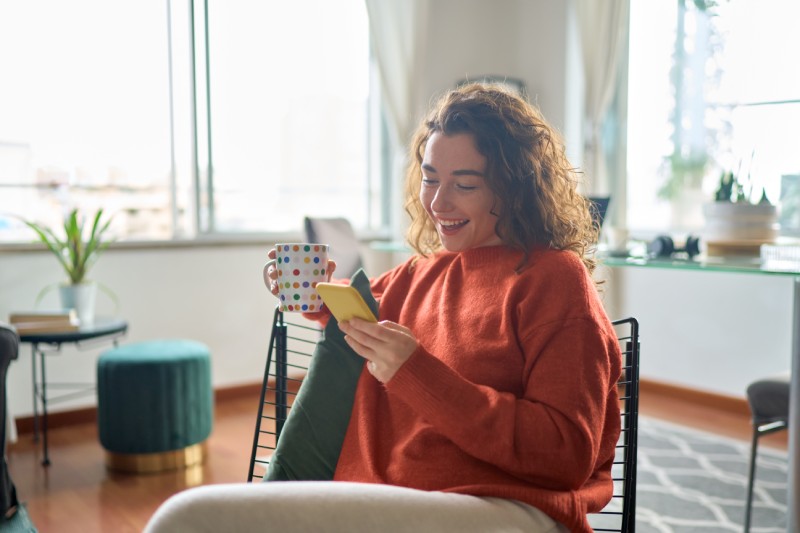 Woman smiling at smartphone while drinking coffee at home